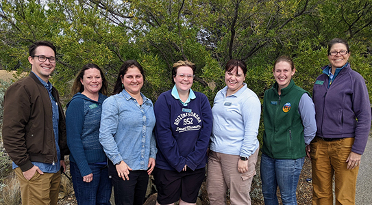 A group of seven Day Camp Teachers standing in front of green shrubbery. The sun is shining and they are all smiling broadly.